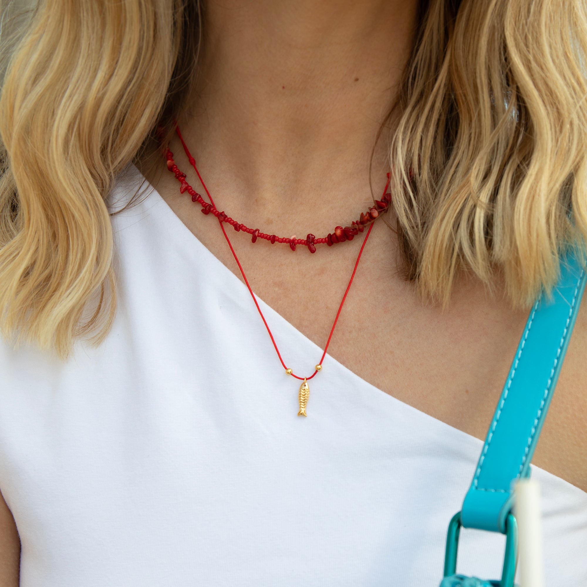 Person wearing a red beaded necklace with a small pendant, white top, and blue strap over shoulder - seolgold