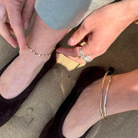 Close-up of a person's feet wearing gold anklets and a silver bracelet on a sandy surface - seolgold