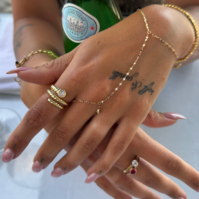 A close-up of hands wearing multiple gold-toned jewelry pieces, including a delicate chain hand bracelet, stacked rings, and a ring with a red gemstone, against a softly blurred background - seolgold