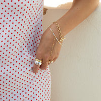 A close-up of a person's hand and wrist wearing delicate gold chain jewelry, including a chain bracelet, chain anklet, and rings, styled with a white dress featuring red polka dots against a light beige wall - seolgold