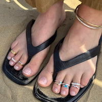 Black sandals worn on feet with multiple rings on toes, on a sandy surface - seolgold