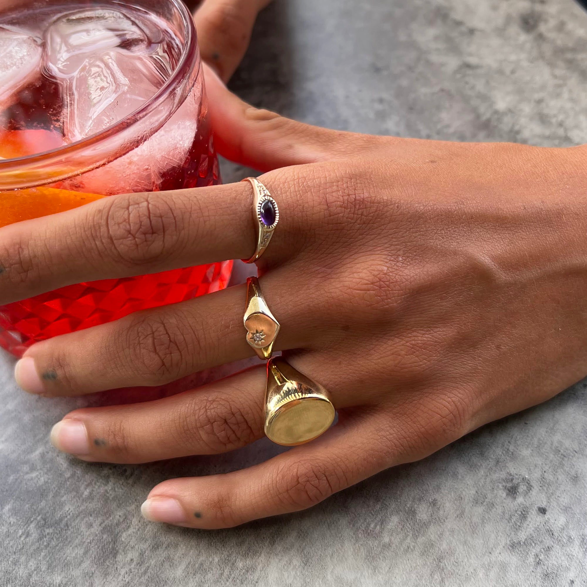 Hand wearing a gold ring with a purple gemstone, holding a red drink - seolgold