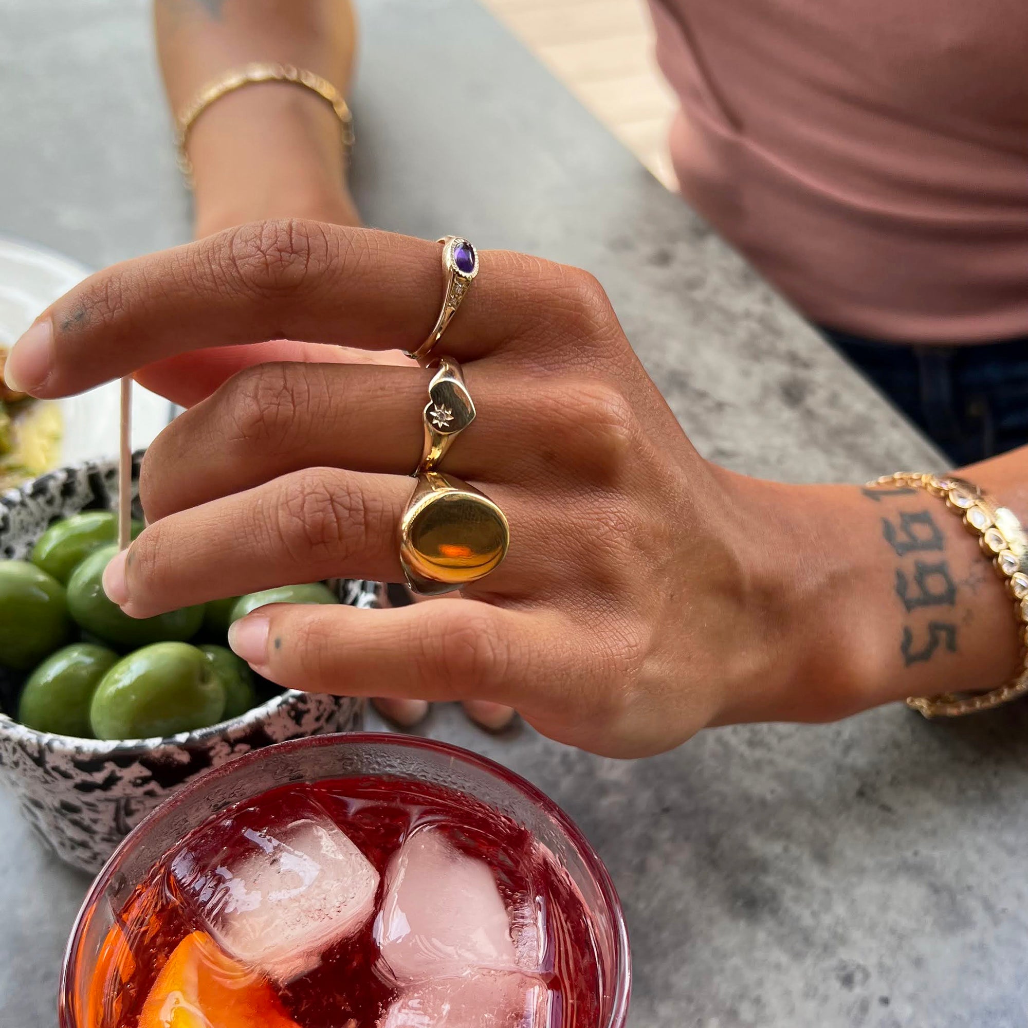 Hand with gold rings and bracelets near a cocktail with ice and lime - seolgold