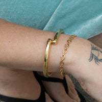 A close-up of a person's wrist wearing two gold-toned bracelets: a thick polished bangle and a delicate chain bracelet, against a turquoise fabric background - Seol gold