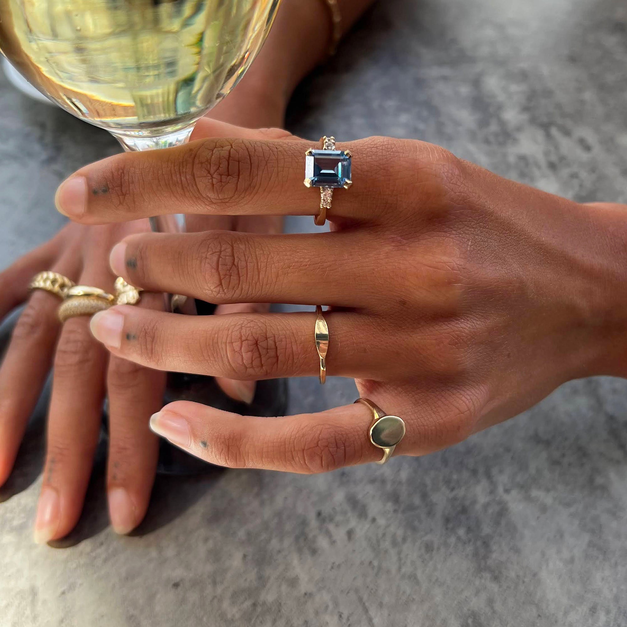 Close-up of hands wearing multiple rings with a glass of white wine in the background - seolgold