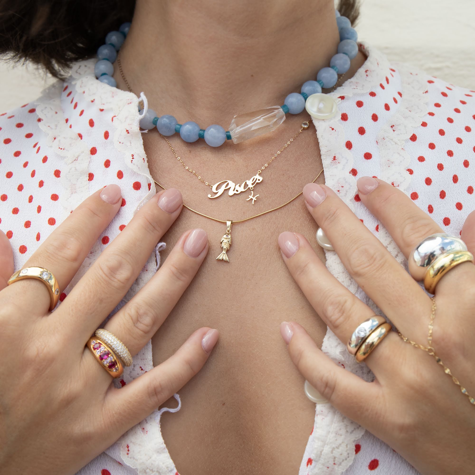 A close-up lifestyle shot of a person wearing layered necklaces and rings. The necklaces include a light blue beaded choker, a gold chain with a 'Pisces' nameplate and a delicate gold chain with a small fish charm - seolgold