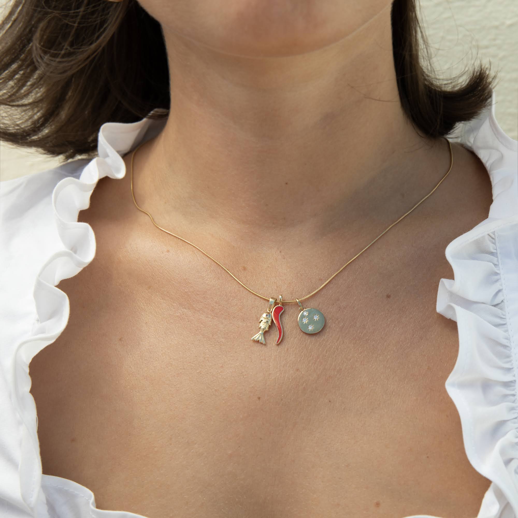 A close-up of a woman's neck and chest wearing a delicate gold necklace with a small ruby fish charm, a gold red cornicello charm, and a round constellation charm - seolgold