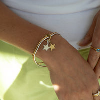 A close-up of a person's wrist wearing a gold bangle bracelet with two dangling star charms, one gold and one silver, over a white top and pale green skirt - seolgold