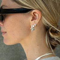A close-up side profile of a woman wearing a pair of silver star-shaped stud earrings, with one larger star and one smaller star, along with black sunglasses and a silver necklace - seolgold