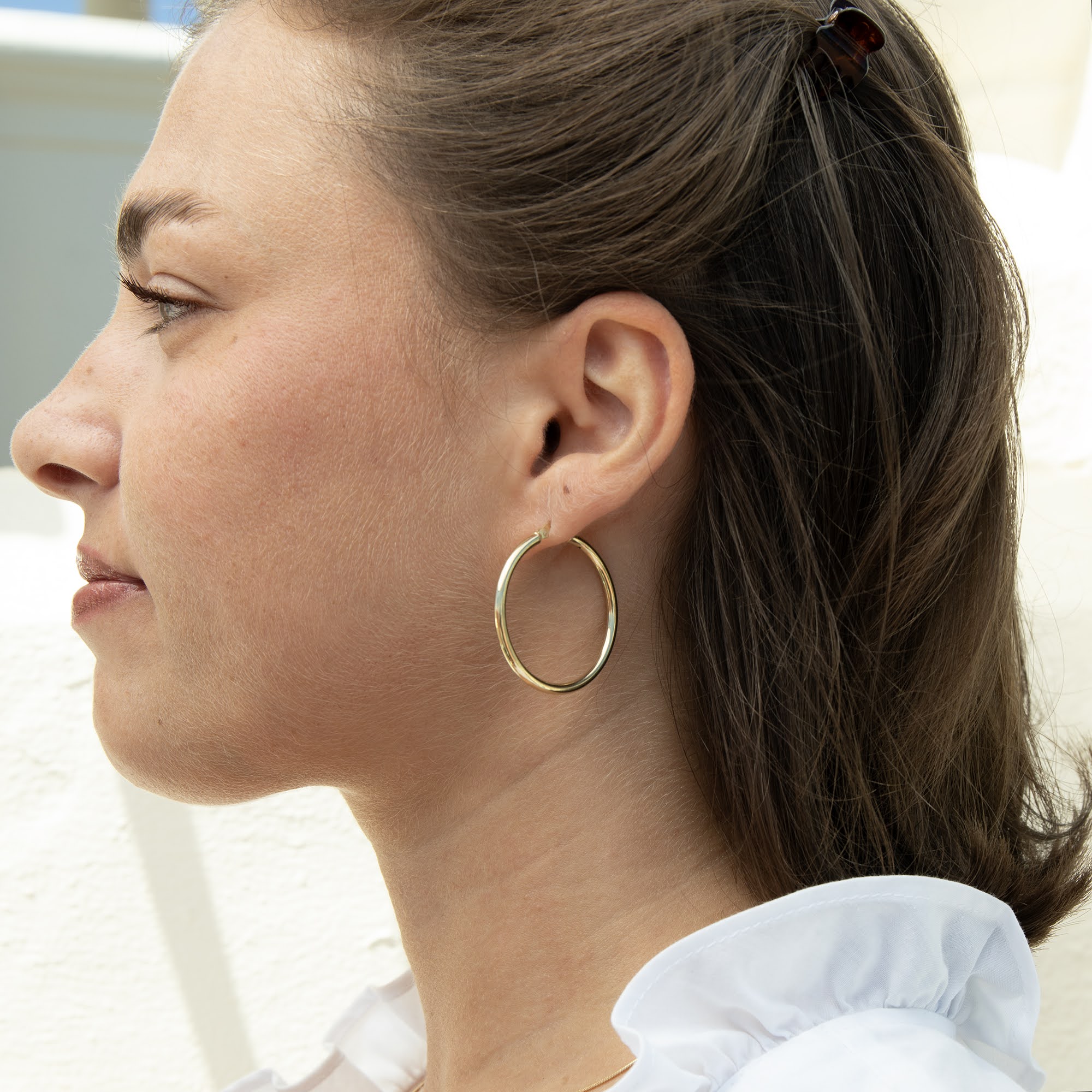 A woman shown in profile wearing a large, thin gold hoop earring. The shot is a close-up of her ear, neck, and side profile, with her brown hair tied back and a white ruffled shirt visible - seolgold