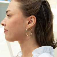 A woman shown in profile wearing a large, thin gold hoop earring. The shot is a close-up of her ear, neck, and side profile, with her brown hair tied back and a white ruffled shirt visible - seolgold
