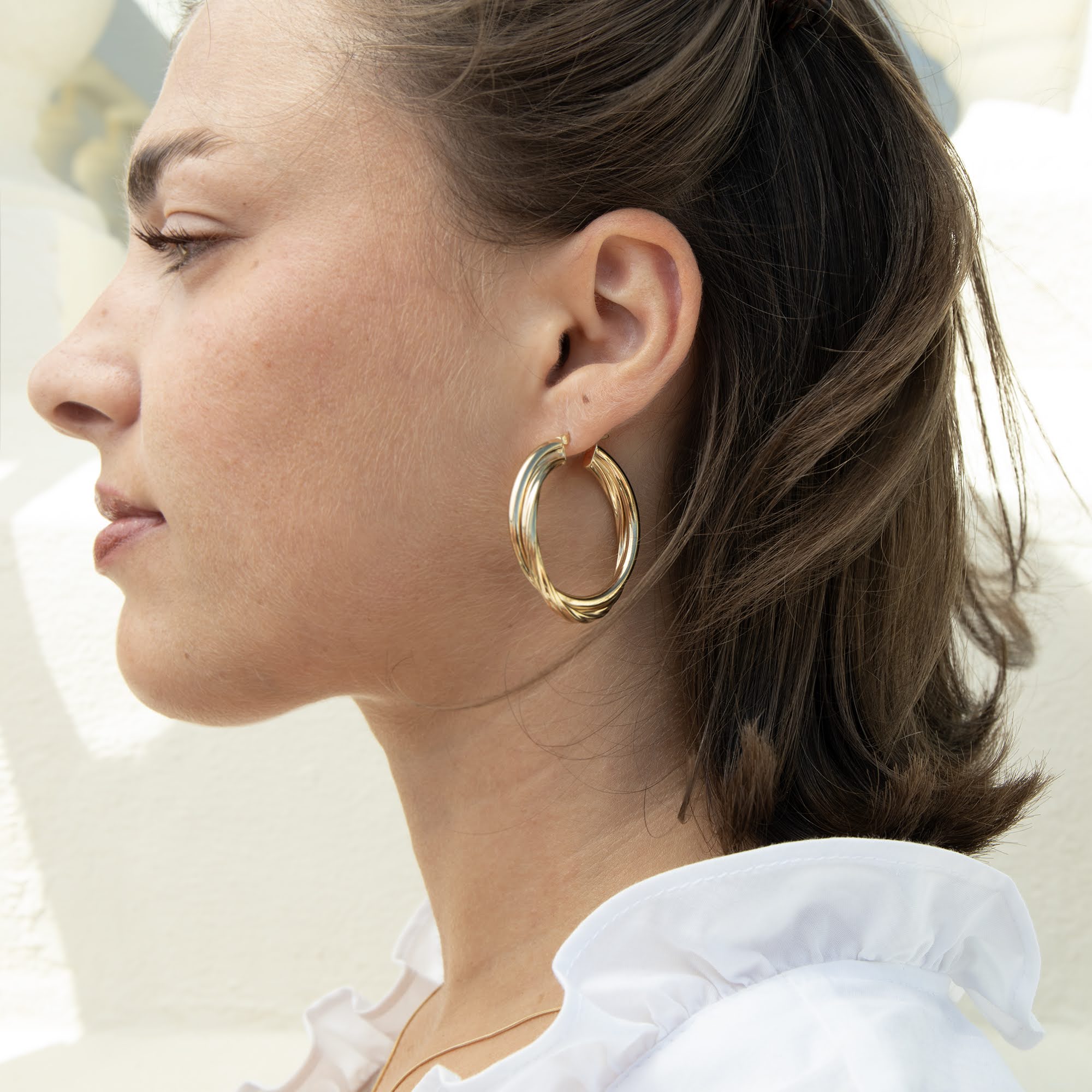 A close-up side profile of a woman wearing a pair of thick, polished gold hoop earrings, with her hair tied back and a white ruffled top visible - seolgold
