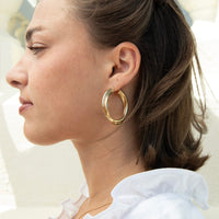 A close-up side profile of a woman wearing a pair of thick, polished gold hoop earrings, with her hair tied back and a white ruffled top visible - seolgold