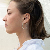 A woman in profile wearing a large, thin silver hoop earring against a light textured wall - seolgold
