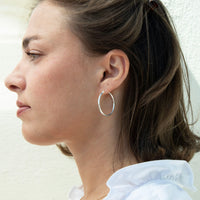 A close-up side profile of a woman wearing a thin, polished silver hoop earring, set against a light textured wall - seolgold