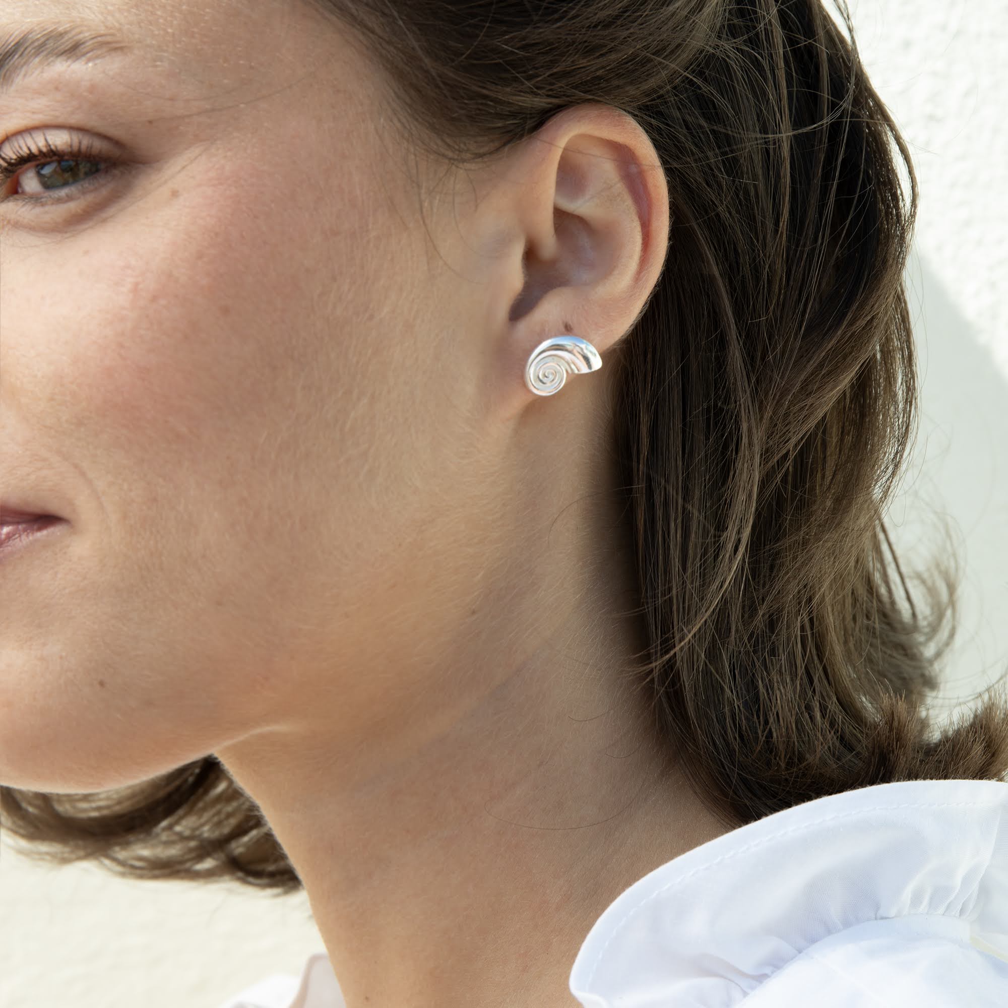 A close-up of a woman's ear wearing a small silver spiral stud earring against a light neutral background - seolgold