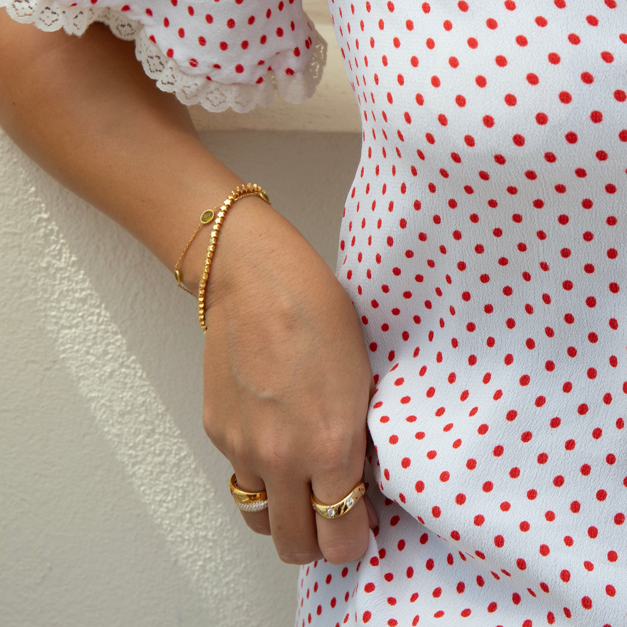 A close-up of a person's hand wearing delicate gold bracelets and rings, next to a white top with red polka dots - seolgold