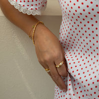 A close-up of a person's hand wearing gold rings and a delicate gold beaded bracelet, paired with a white dress featuring red polka dots and a lace-trimmed sleeve, set against a neutral textured wall - seolgold
