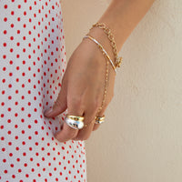 A close-up of a person's hand and wrist wearing a delicate gold chain bracelet with a small charm, layered with a thin silver chain and two gold rings. The person is wearing a white dress with red polka dots against a light beige wall - seolgold