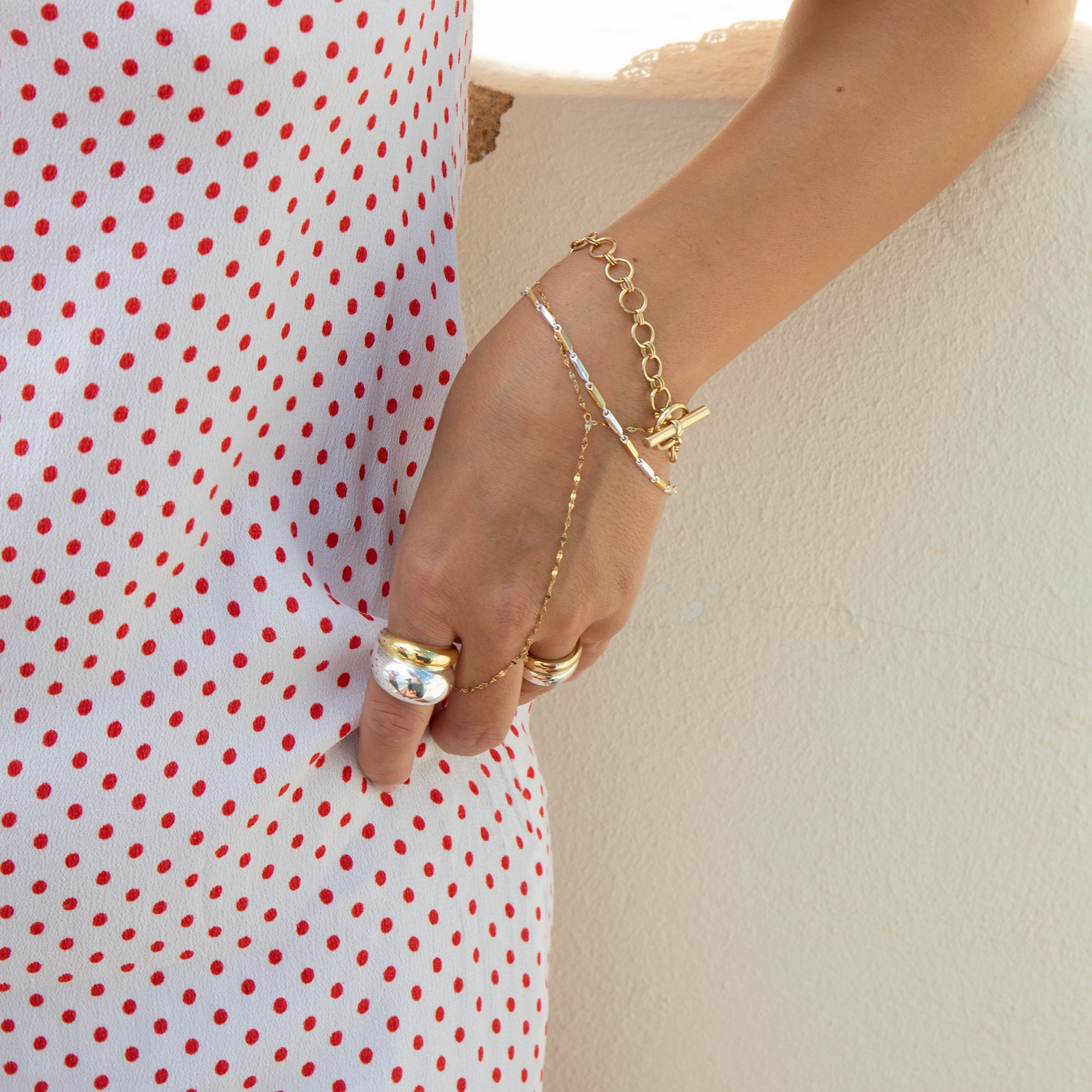 A close-up of a person's hand and wrist wearing delicate gold chain jewelry, including a chain bracelet, chain anklet, and rings, styled with a white dress featuring red polka dots against a light beige wall - seolgold