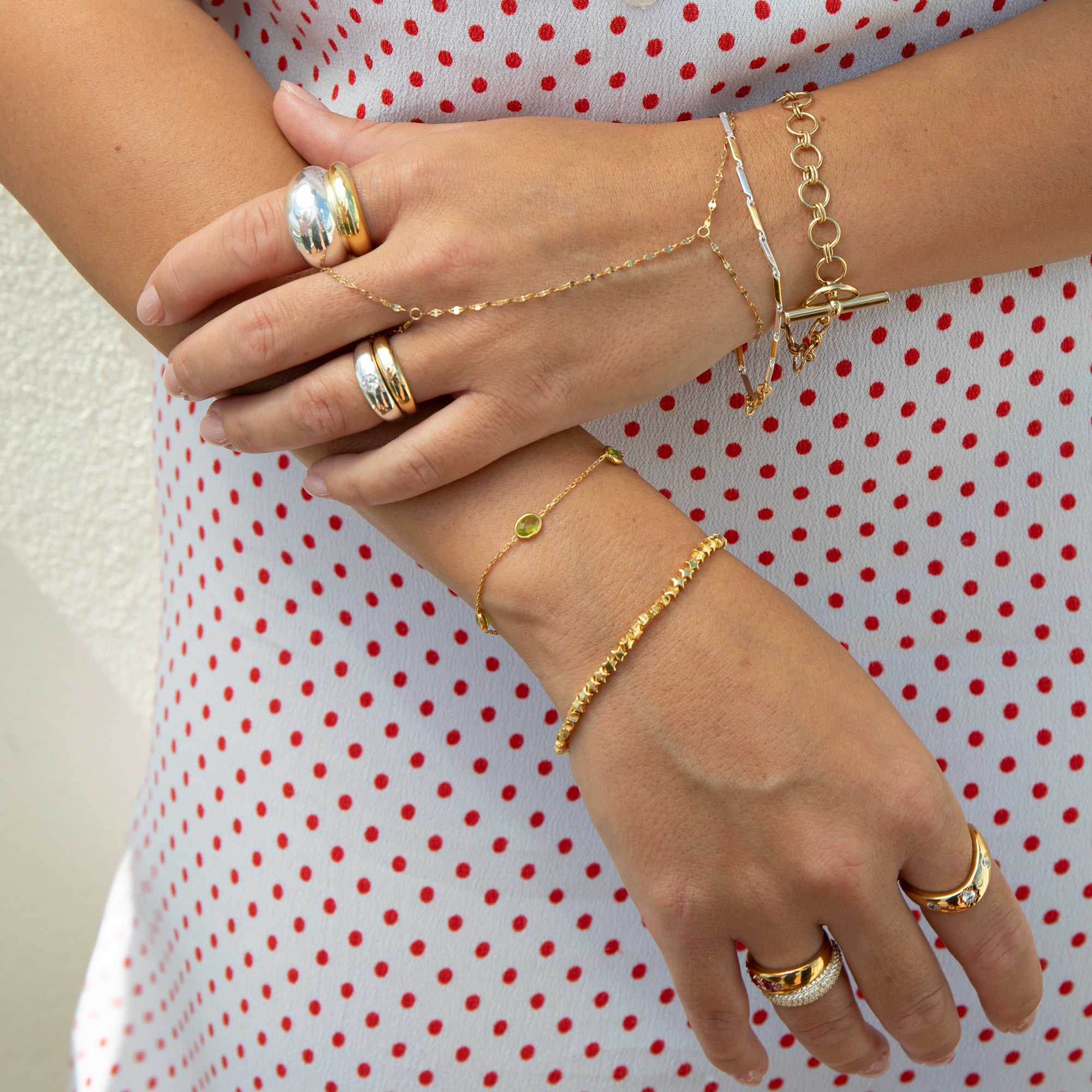 A close-up of a person's hands and arms wearing multiple delicate gold bracelets and rings, including chain bracelets, beaded bracelets, and stacked rings, styled with a white dress featuring red polka dots - seolgold