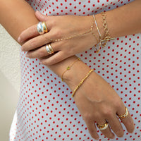 A close-up of a person's hands and arms wearing multiple delicate gold bracelets and rings, including chain bracelets, beaded bracelets, and stacked rings, styled with a white dress featuring red polka dots - seolgold