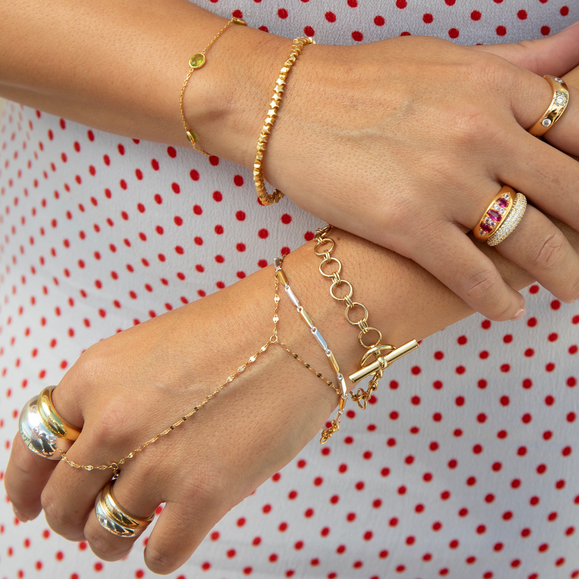 Gold jewelry set on a model's hands, including a delicate chain bracelet with a toggle clasp, a beaded bracelet, a ring with pink gemstones, and a gold signet ring, against a white and red polka dot background - seolgold