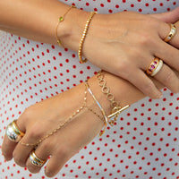 Gold jewelry set on a model's hands, including a delicate chain bracelet with a toggle clasp, a beaded bracelet, a ring with pink gemstones, and a gold signet ring, against a white and red polka dot background - seolgold