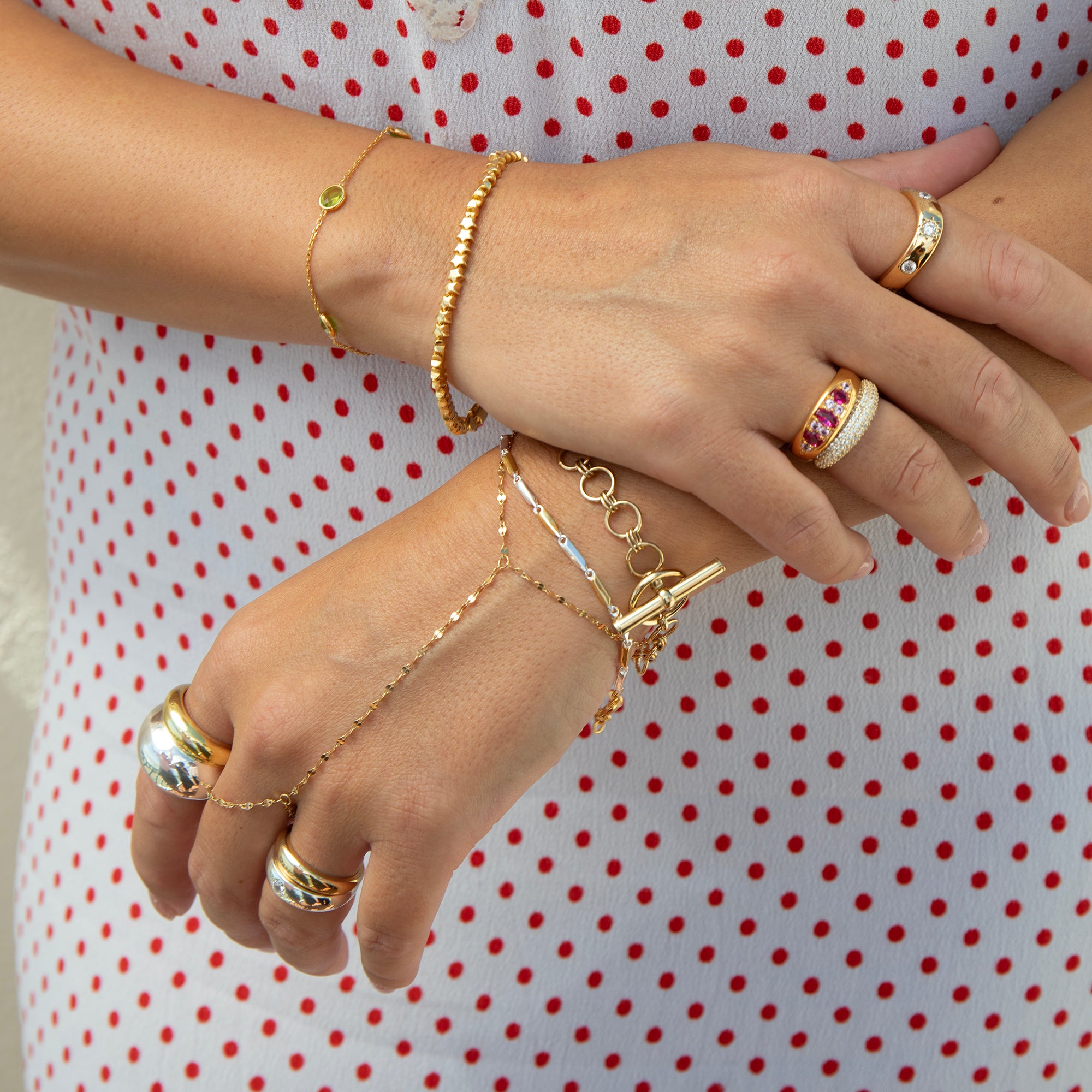 A close-up of a person's hands and arms wearing multiple gold-toned jewelry pieces, including bracelets, rings, and a delicate chain hand bracelet, against a white dress with red polka dots - seolgold