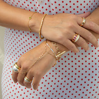 A close-up of a person's hands and arms wearing multiple gold-toned jewelry pieces, including bracelets, rings, and a delicate chain hand bracelet, against a white dress with red polka dots - seolgold