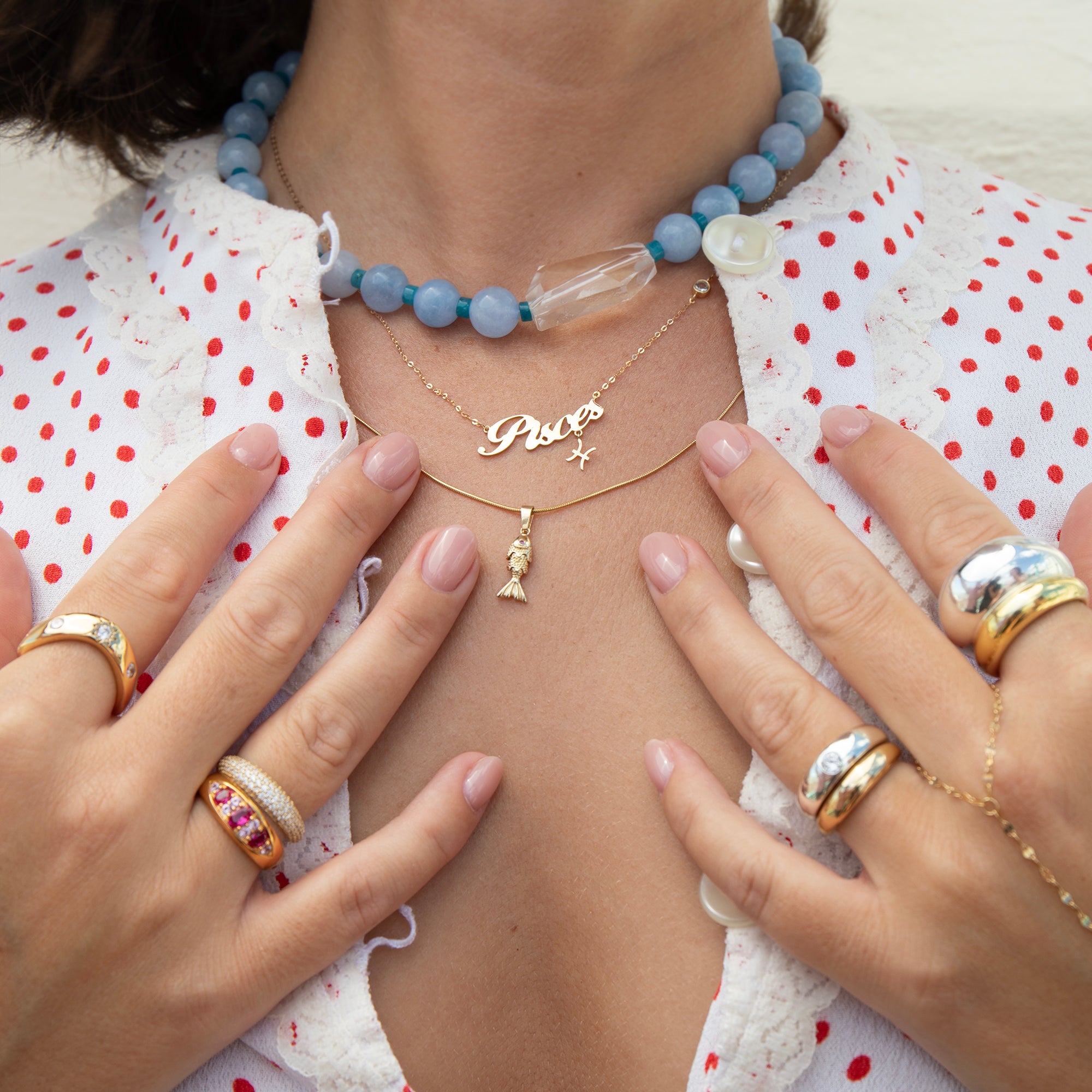 A close-up lifestyle shot of a person wearing layered jewelry, including a light blue beaded choker, a gold nameplate necklace reading 'Pico' with a mermaid charm, and multiple gold rings on both hands, over a white lace top with red polka dots - seolgold