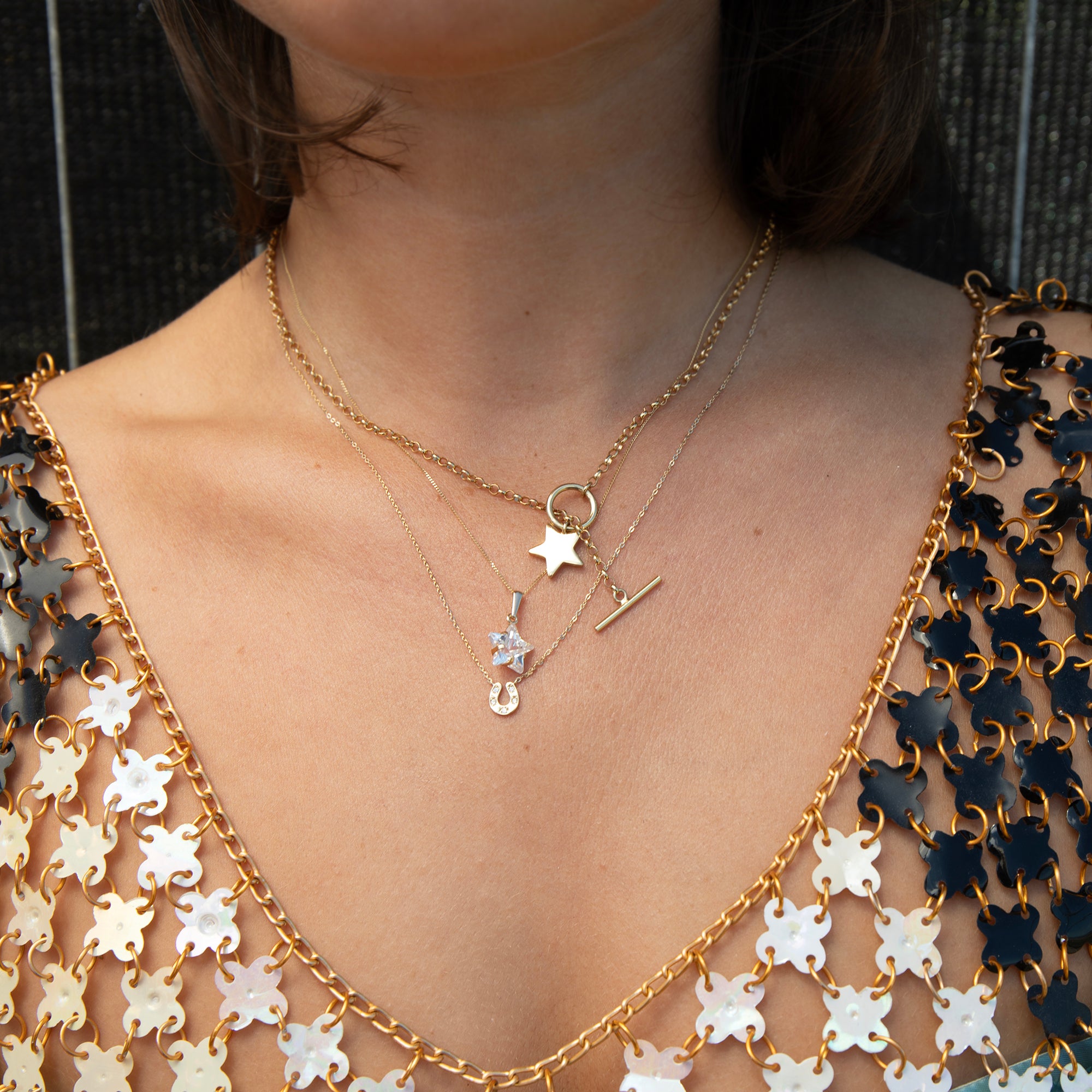 A close-up of a person's neck and chest wearing layered gold necklaces, including a star charm, a horseshoe charm, and a small cross charm, over a black and white patterned top - seolgold