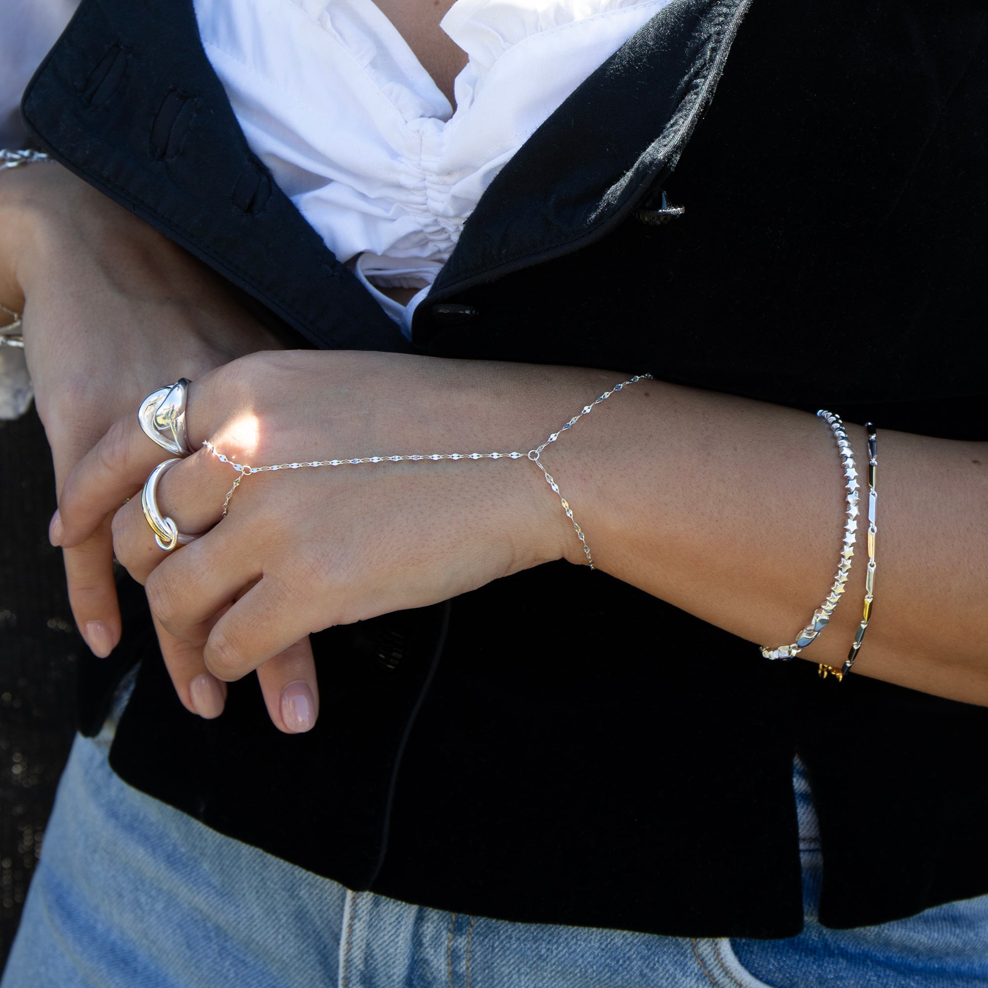 A close-up of a person's hands and arms wearing a delicate silver Y-shaped chain hand bracelet, paired with gold rings and silver bangles, styled with a white blouse and black blazer - seolgold