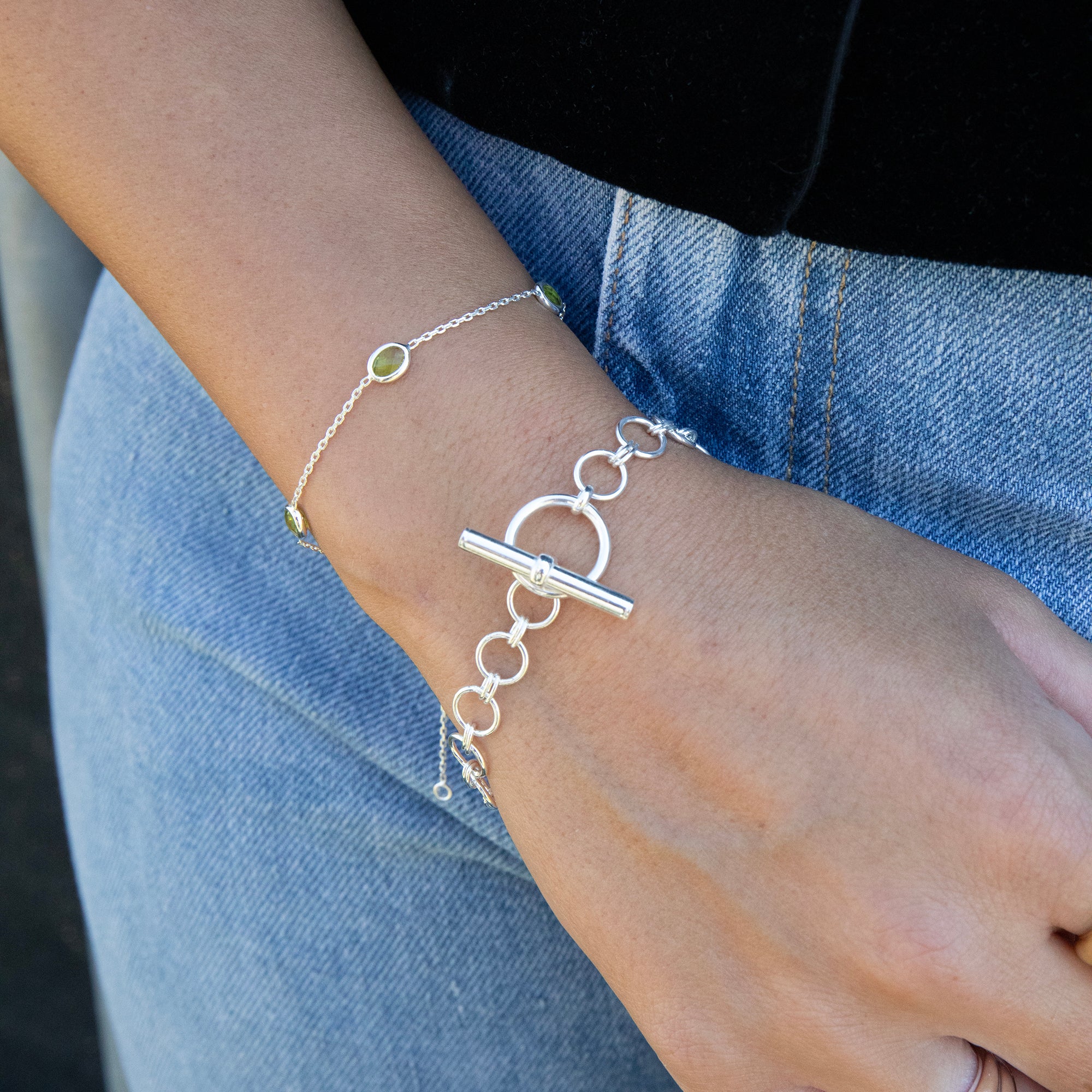 A close-up of a person's wrist wearing two delicate bracelets: a silver chain bracelet with a large toggle clasp and a silver link bracelet with a small green stone accent, paired with blue denim jeans - seolgold