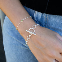 A close-up of a person's wrist wearing two delicate bracelets: a silver chain bracelet with a large toggle clasp and a silver link bracelet with a small green stone accent, paired with blue denim jeans - seolgold
