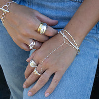 A close-up of a person's hands and lap wearing a delicate silver chain hand bracelet with a matching chain bracelet on the wrist, styled with multiple statement rings - seolgold