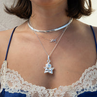 A close-up of a person wearing layered silver necklaces, including a choker, a delicate chain with a small initial pendant, and a longer chain with a cherub pendant, styled with a lace bra top - seolgold