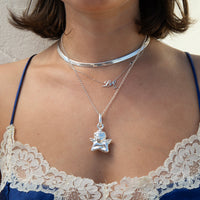 A close-up of a person wearing layered silver necklaces, including a choker, a delicate chain with a small initial pendant, and a longer chain with a cherub pendant, styled with a lace bra top - seolgold
