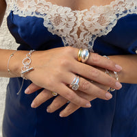 A close-up of a person's hands wearing multiple silver rings and bracelets, with a navy blue satin dress featuring a white lace bodice in the background - seolgold