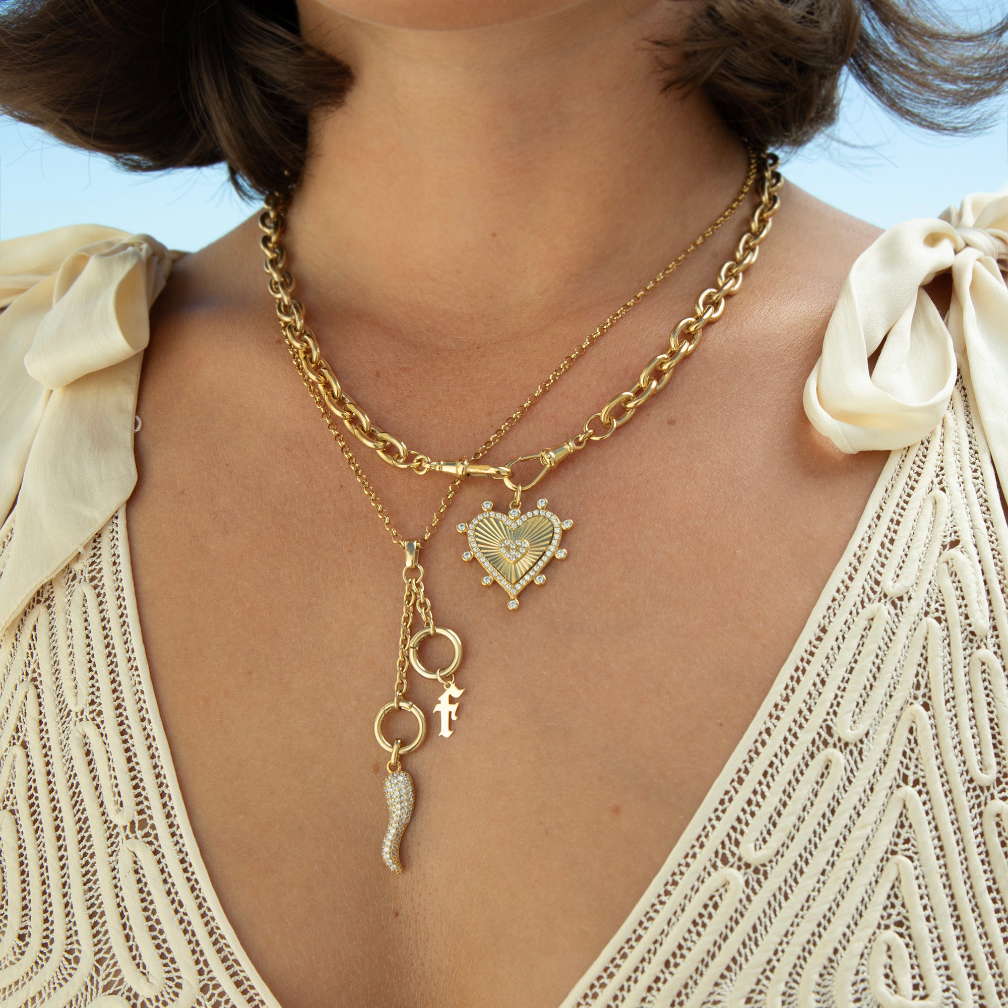 A close-up of a person wearing layered gold necklaces, including a curb chain, a delicate chain with a heart-shaped pendant, and dangling charms, paired with a cream lace top - seolgold