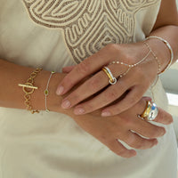 A close-up of a person's hands and arms wearing multiple delicate jewelry pieces, including a gold toggle bracelet, a thin green bead bracelet, a silver-and-gold ring, and a chain bracelet with a large silver ring on the middle finger, styled with a cream-colored embroidered top - Seolgold