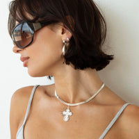 A woman in profile wearing a silver cross pendant necklace, silver hoop earrings, and dark sunglasses against a plain light wall - seolgold