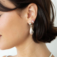 A close-up of a woman's ear wearing a silver teardrop dangle earring with a small silver stud earring, set against a clean white background - seolgold