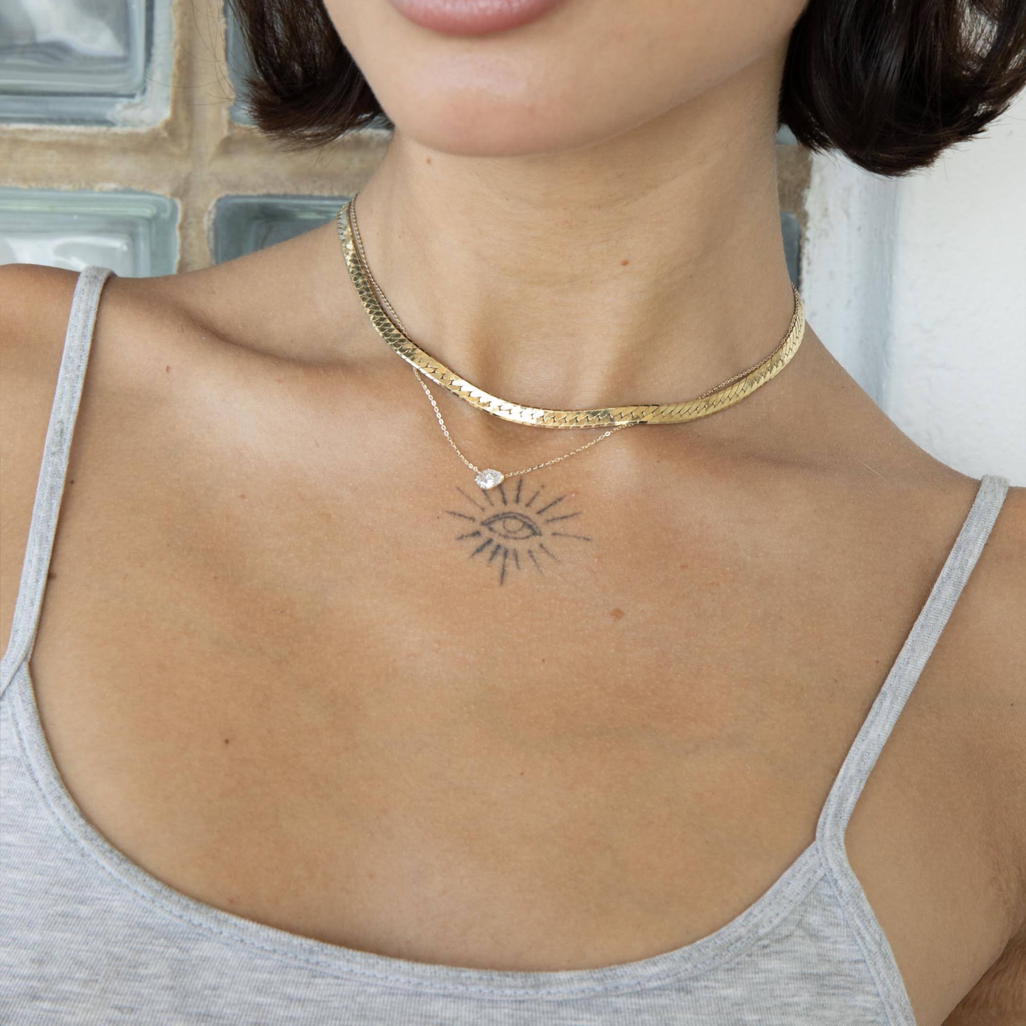 A close-up of a woman's neck and upper chest wearing a layered gold necklace. The top layer is a textured choker, and the bottom layer is a delicate chain with a small eye pendant and a clear heart-shaped stone - seolgold