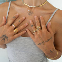 A close-up lifestyle shot of a model wearing multiple gold jewelry pieces, including a choker necklace, layered necklaces, rings, a chain bracelet, and a hand chain, styled with black tattoos on her chest and hands - seolgold
