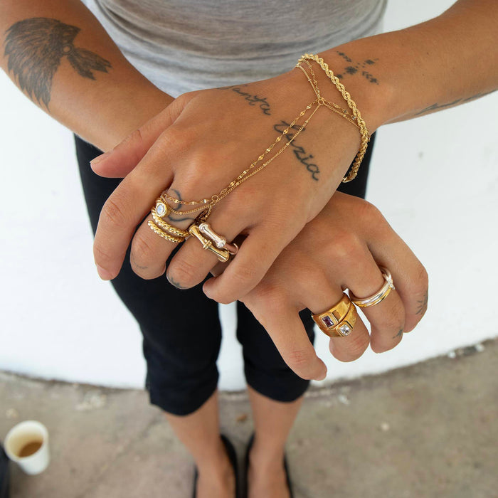 A close-up of a person's hands and arms wearing multiple gold rings, a gold chain bracelet, and a gold chain hand warmer, paired with a gray t-shirt and black pants - seolgold