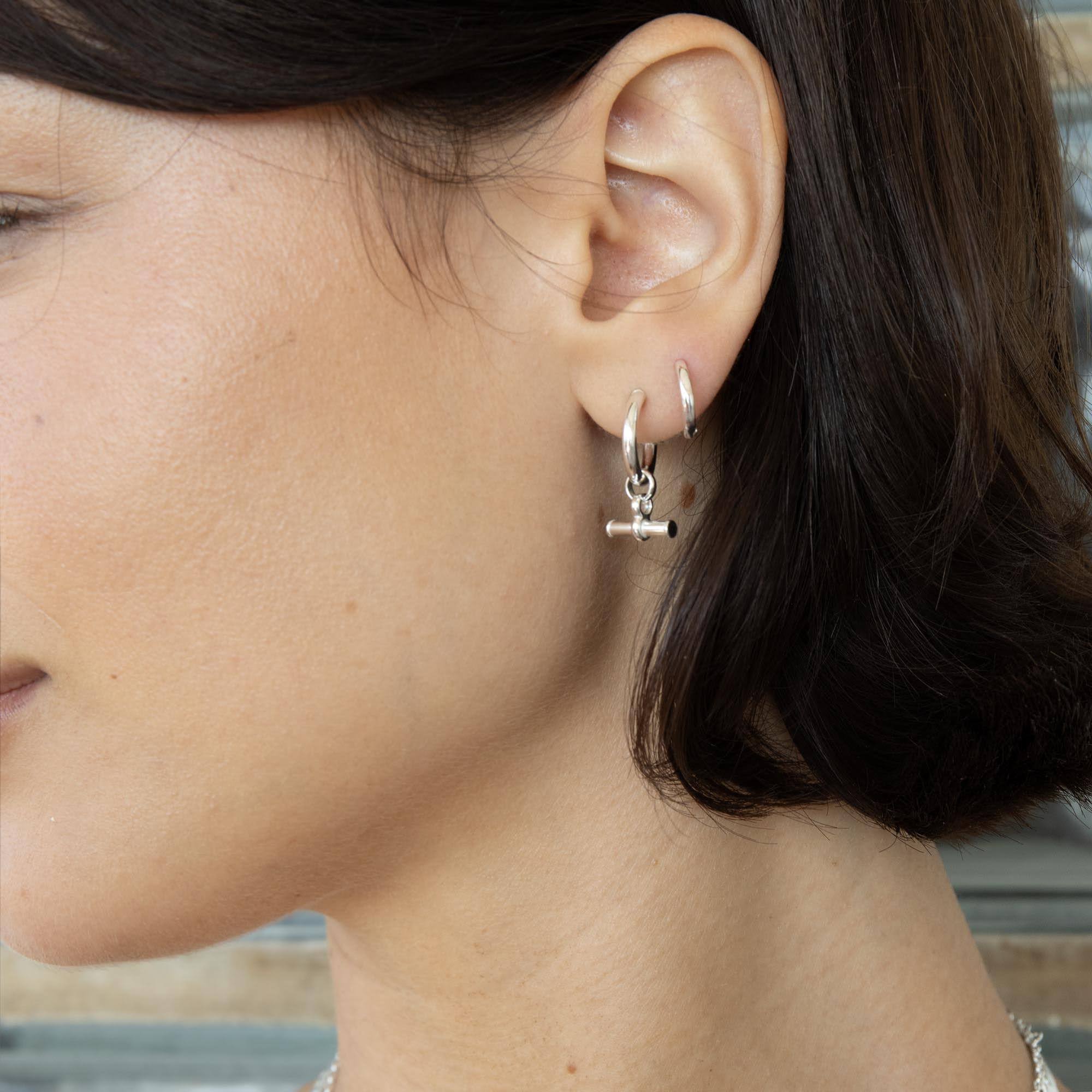 A close-up side profile of a woman wearing a small silver huggie hoop earring with a dangling toggle t-bar charm on her earlobe - seolgold