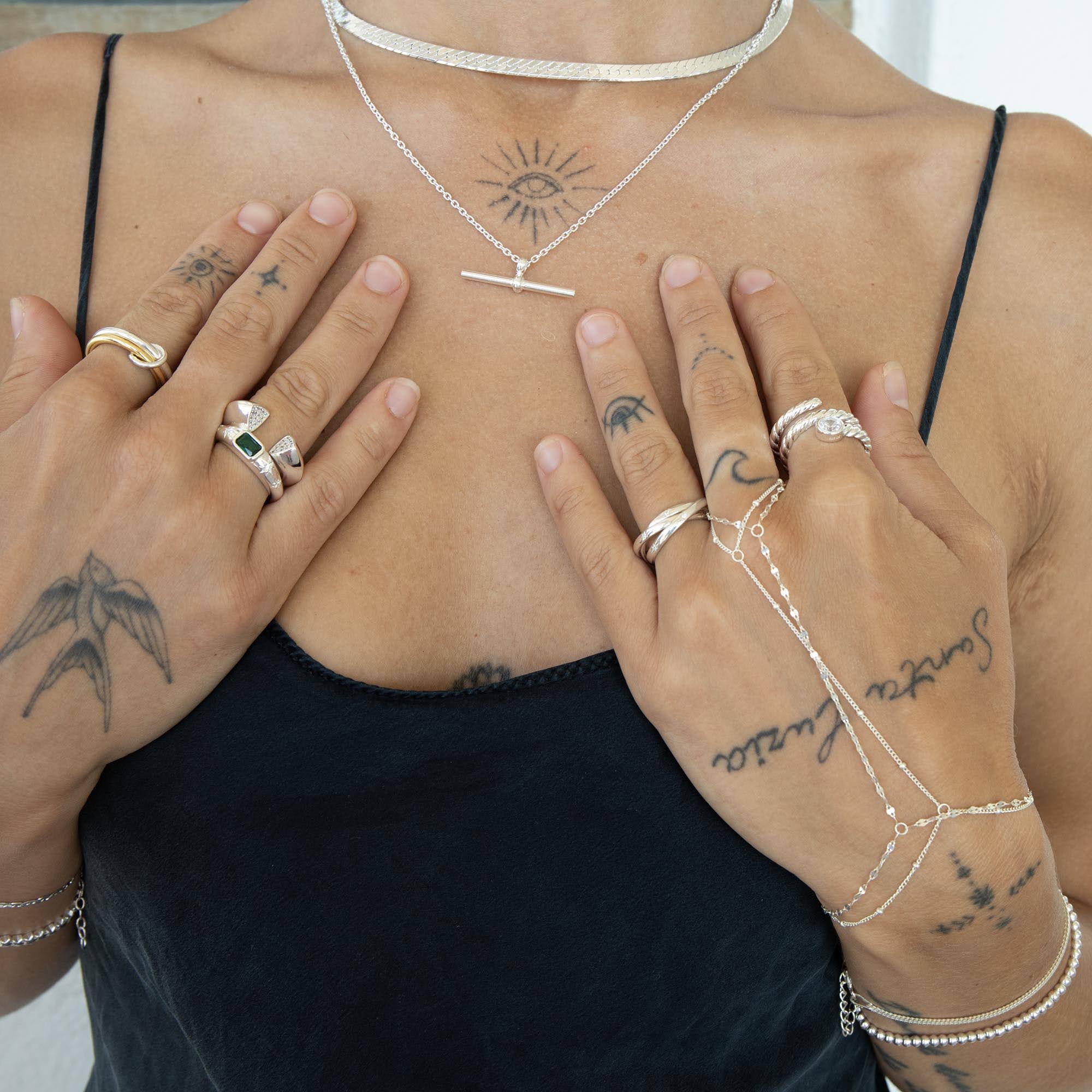 A close-up of a person's chest and hands wearing multiple delicate silver jewelry pieces, including a necklace, rings, and chain bracelets, over a black camisole top - seolgold