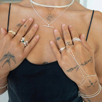 A close-up of a person's chest and hands wearing multiple delicate silver jewelry pieces, including a necklace, rings, and chain bracelets, over a black camisole top - seolgold