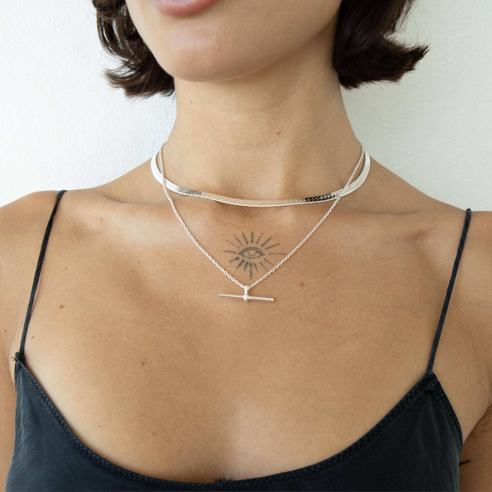 A close-up of a model's neck and upper chest wearing a layered silver necklace set. The set includes a wide, hammered band choker and a delicate chain with a T-bar pendant - seolgold